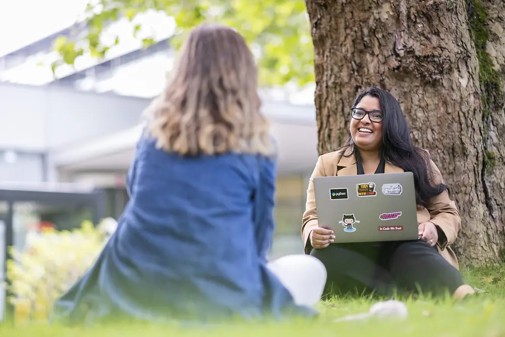 Zwei Frauen unterhalten sich sitzend auf einer Wiese, dabei hält eine Frau einen Laptop in der Hand.