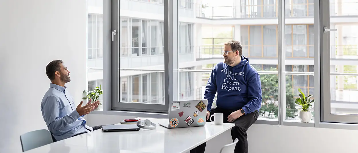 Zwei junge Männer in einem hellen Bürogebäude unterhalten sich entspannt.