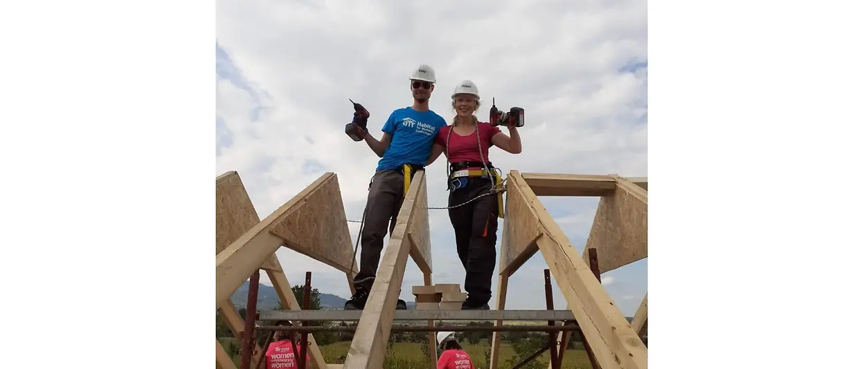 Zwei Helfer:innen stehen auf dem Holzgerüst und posieren für das Foto, indem sie ihre Bohrmaschinen in die Höhe halten.