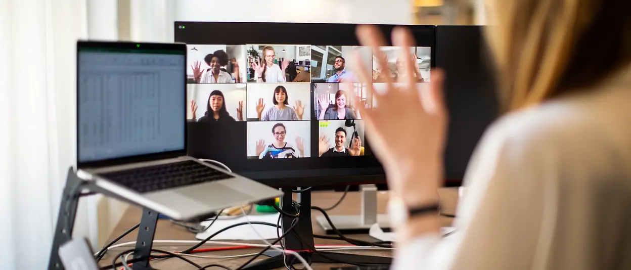 Eine Frau sitzt vor ihrem Computer und winkt ihren Kolleg:innen zu, die sie bei einem Online-Meeting auf dem Computerbildschirm sieht. 