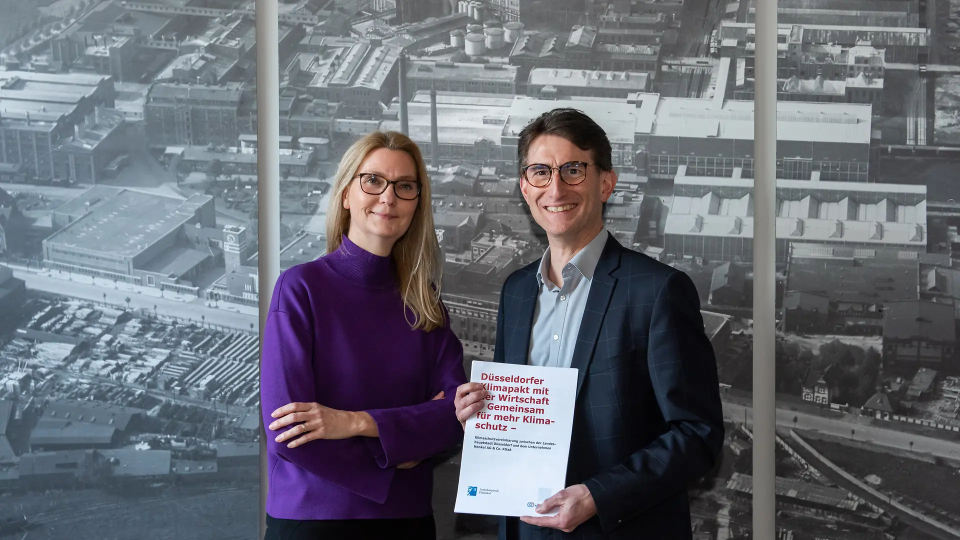 Eine Frau und ein Mannstehen nebeneinander und lächeln in die Kamera. Der Mann hält ein Papier mit dem Titel des Klimapaktes.