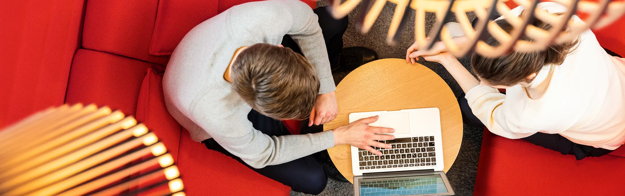 Zwei Personen sitzen auf roten Sofas und arbeiten zusammen an einem Laptop