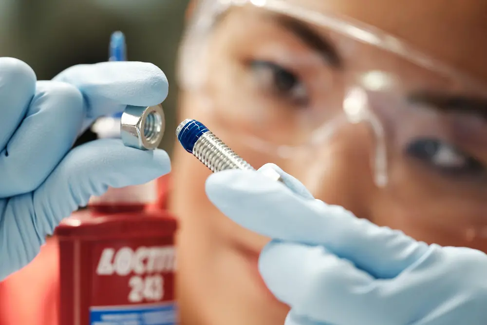 Close-up of a person with safety glassess. They have a bolt in one hand, and in the other hand a screw on which a blue LOCTITE threadlocker has been applied. The threadlocker bottle can be partially seen.