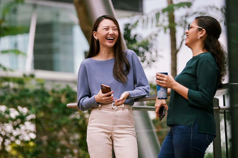 Zwei Frauen stehen im begrünten Foyer eines modernen Bürogebäudes und lachen.