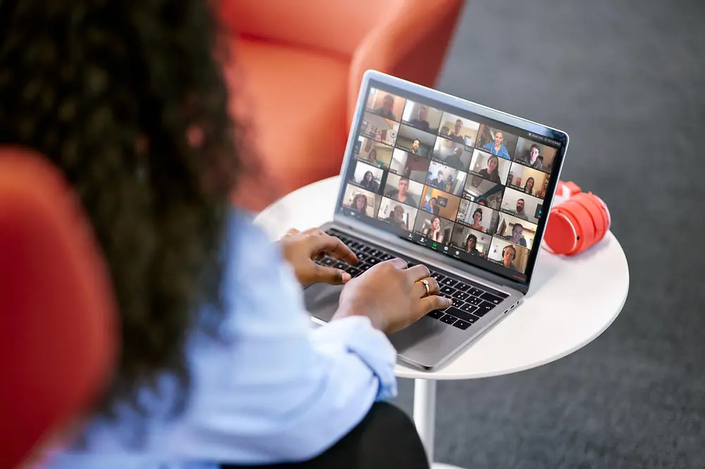 Eine Frau mit langen braunen Haaren sitzt an einem Tisch mit dem Rücken zur Kamera. Auf dem Tisch vor ihr steht ihr Laptop, auf dem eine virtuelle Besprechung mit Kolleg:innen zu sehen ist.