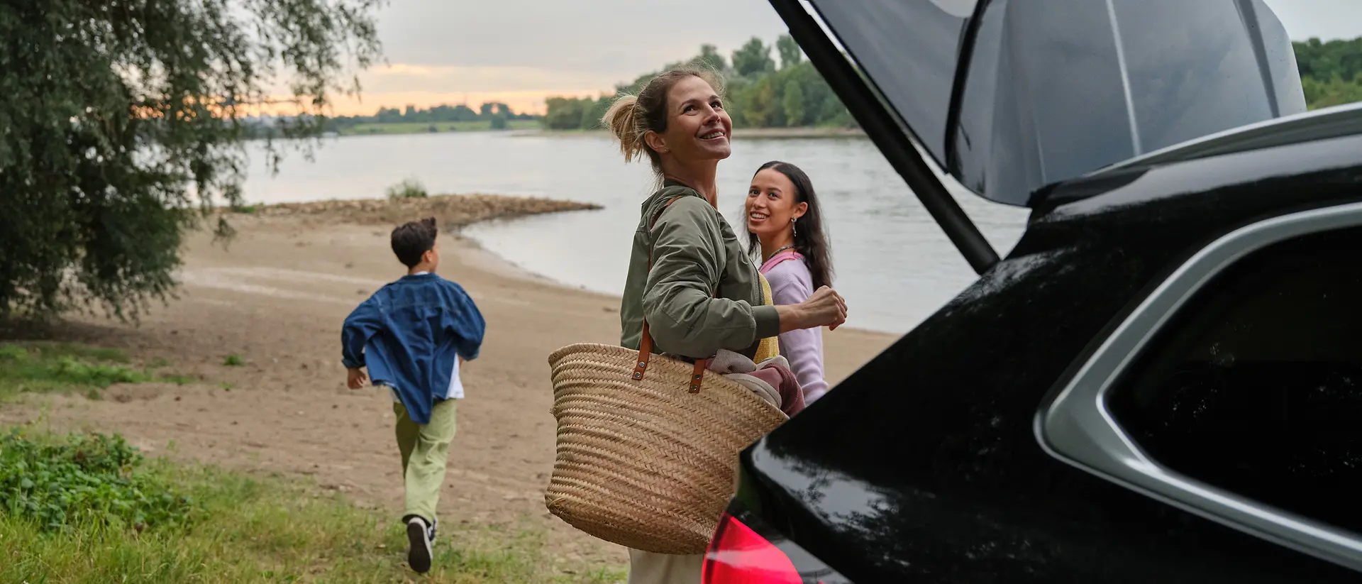 Eine Mutter ist gerade dabei den Kofferraum zu schließen während ihre Tochter ihr zuschaut und der Sohn schon am Strand auf das Wasser zuläuft.