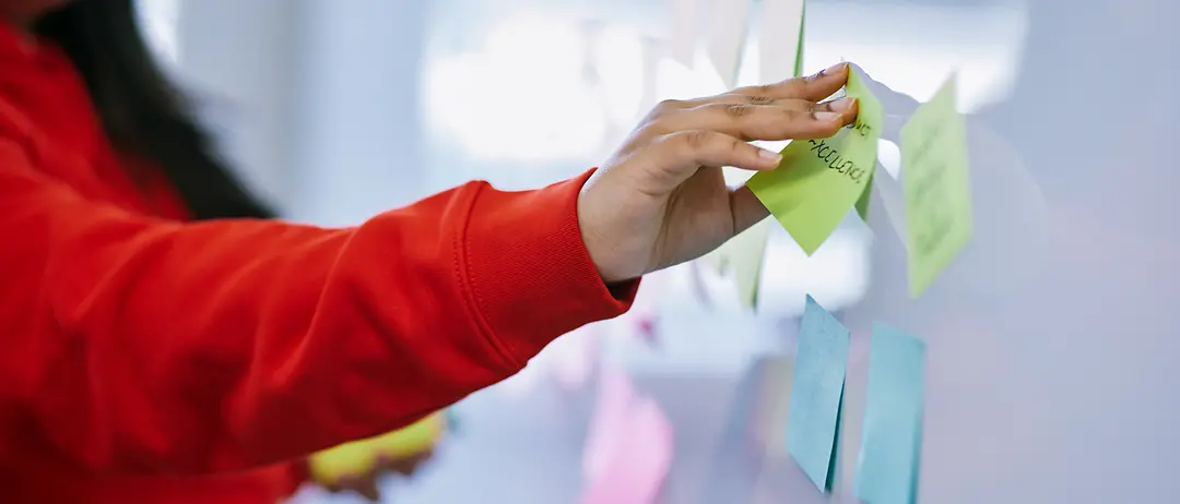 Eine Frau in einem roten Pullover klebt Post-Its an ein Whiteboard.