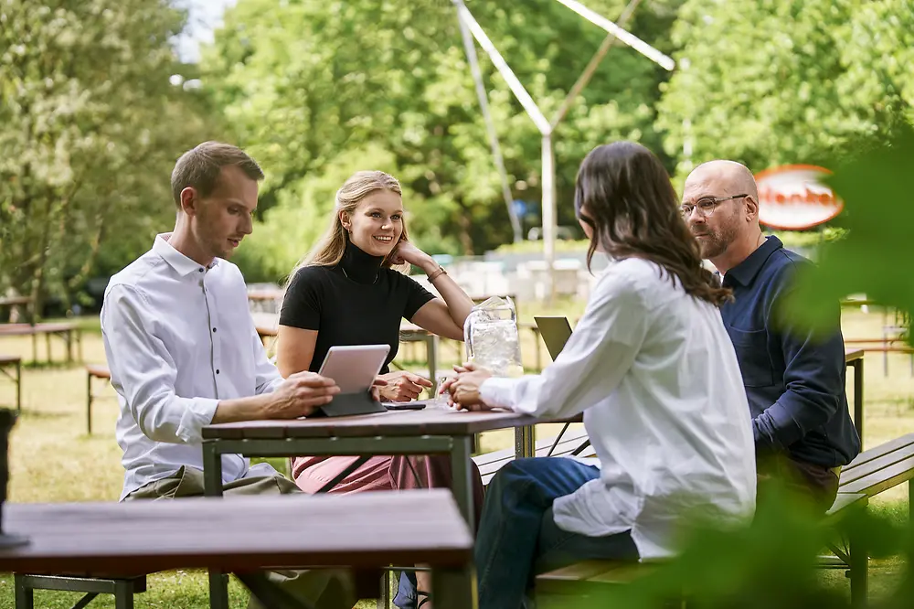 Eine Gruppe von vier Personen sitzt draußen mit einem Laptop und einem Tablet an einem Tisch und unterhält sich. Im Hintergrund sind Bäume und eine sonnige Umgebung zu sehen.