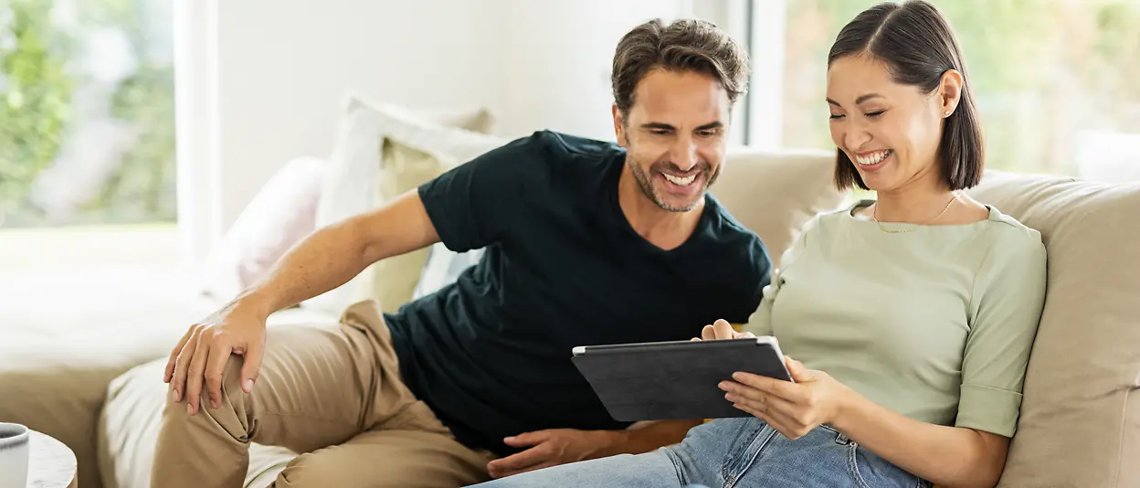 Ein Mann und eine Frau sitzen zu Hause auf ihrem Sofa und schauen gemeinsam auf ein Tablet.