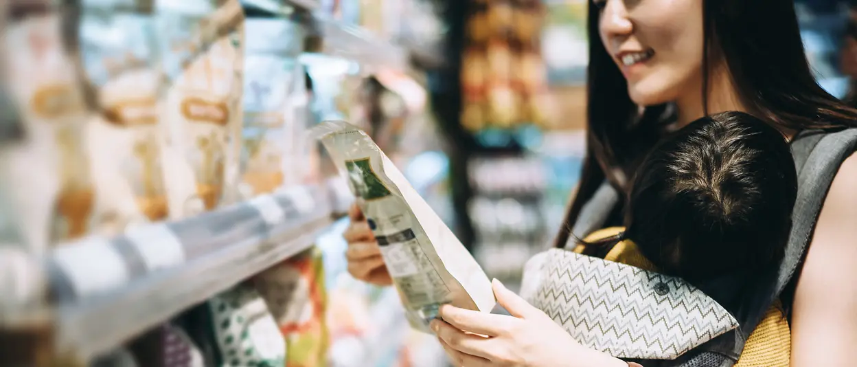 
Eine Frau mit ihrem Baby steht vor einem Regal in einem Supermarktgang und hält einen in einem Plastikbeutel verpackten Artikel in der Hand.